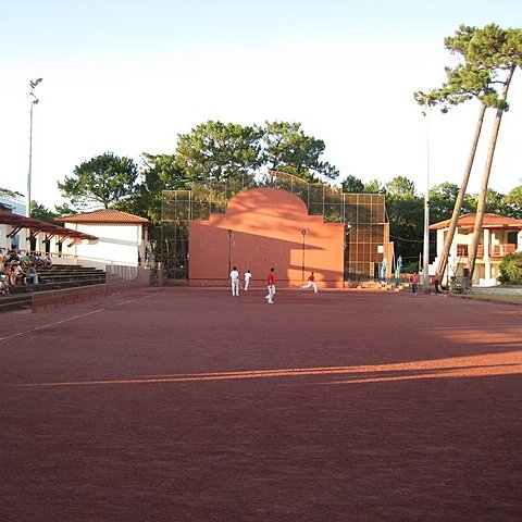Joueurs de pelote basque sur un fronton rouge en plein air, entouré de pins.