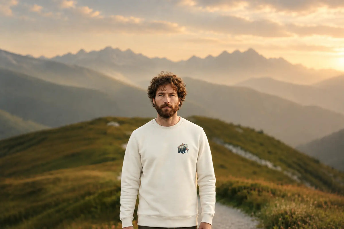 Homme portant un sweat ivoire à col rond avec un motif ours sur la poitrine, devant des montagnes au coucher du soleil.