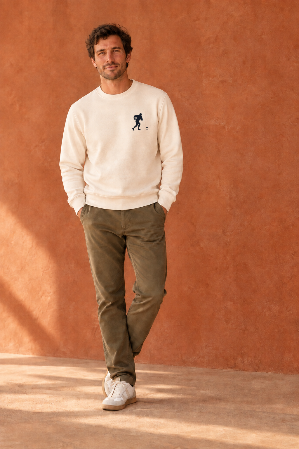 homme les mains dans les poches devant mur etrracota, portant un sweat couleur ivoire au design Rgby du sud-ouest