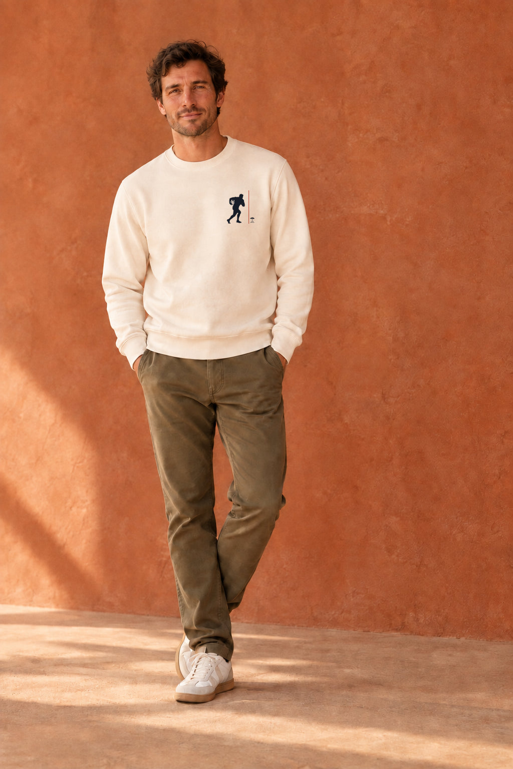 homme debout devant mur terracota, porte un sweat couleur ivoir avec design Rugby
