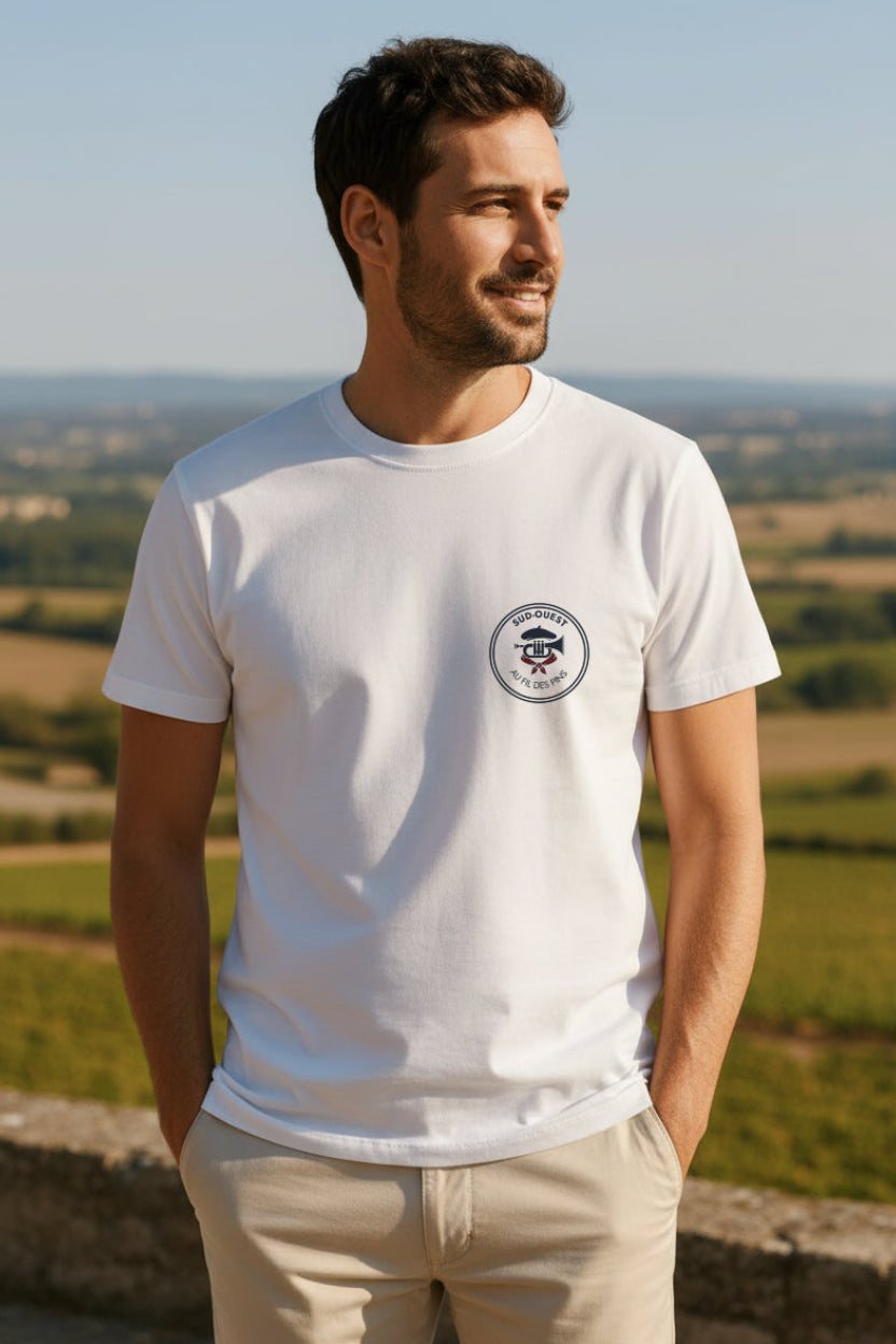homme debout les mains dans les poches devant paysage du Gers, porte un t-shirt blanc au tissu épais avec design des fêtes du sud-ouest